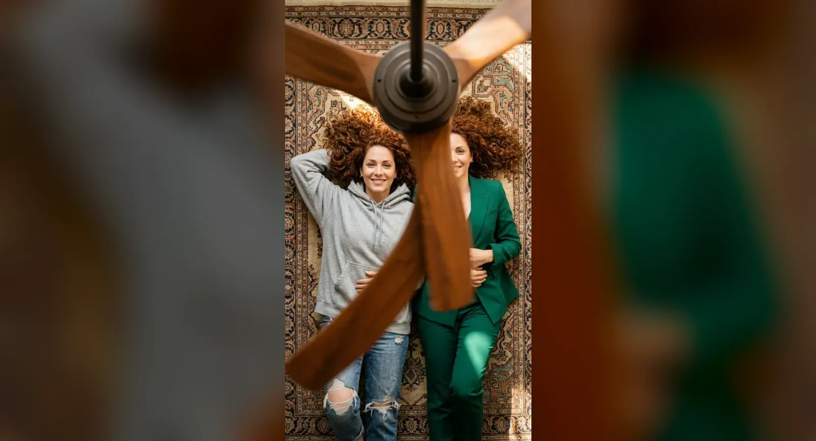 Lena, one of eroq's fictional characters, seen twice from above on a Persian rug under a wooden ceiling fan: in a grey hoodie and jeans on one side of the blade, in an emerald suit on the other.