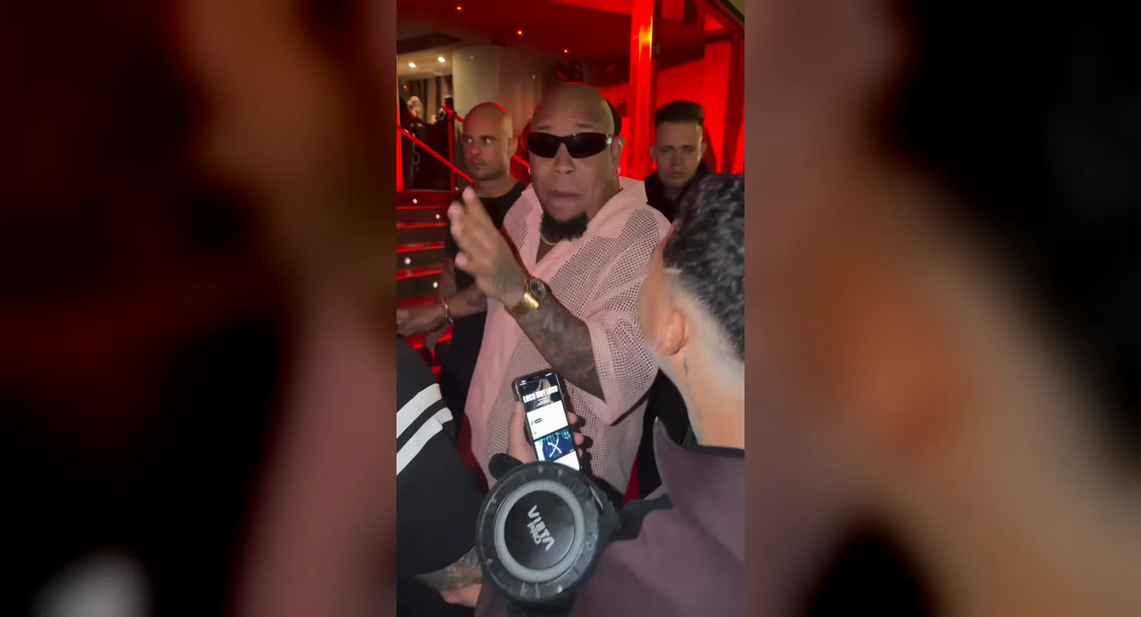 El clip original del freestyle de Bulin 47 grabado con un móvil: el rapero, con gafas de sol negras y camisa rosa de malla, rapea en la entrada de un club iluminada en rojo mientras un fan con un altavoz escucha en primer plano.